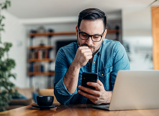 A focused man in glasses uses a smartphone at a wooden table with a laptop and a coffee cup in a modern home office setting.