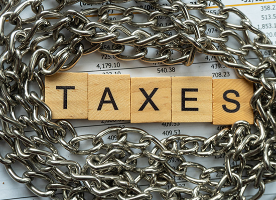 The word 'TAXES' spelled out in scrabble tiles, encircled by a heavy metal chain on top of financial documents, symbolizing the burdensome nature of taxes.