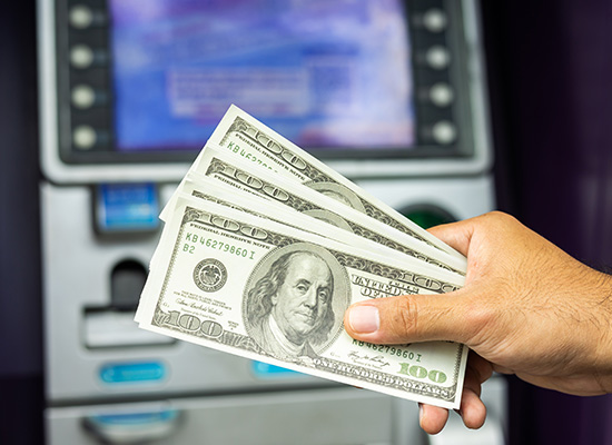 A person's hand holding three hundred-dollar bills in front of an ATM, with the screen displaying transaction options in the background.