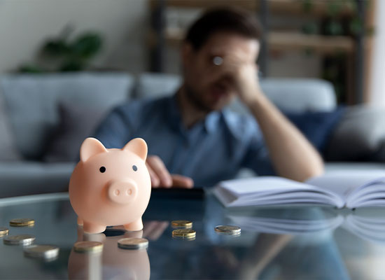 A worried person sits on a couch with their hand on their forehead, looking at an open notebook on a glass table. In the foreground, there is a pink piggy bank and scattered coins—symbolizing savings for retirement income. The background is slightly blurred with shelves and plants.