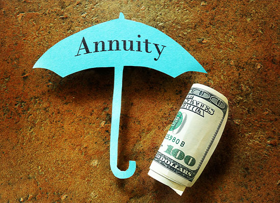 A blue paper cutout in the shape of an umbrella with the word 'Annuity' printed on it lies on a brown surface beside a rolled-up U.S. hundred-dollar bill. The image symbolizes financial protection and retirement income security provided by annuities.
