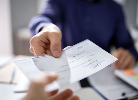 Close-up of a person's hand in a blue shirt giving a check to another person whose hand is slightly out of focus in the foreground. The scene appears to take place in an office setting, with estate planning documents and a pen visible on the desk in the background.