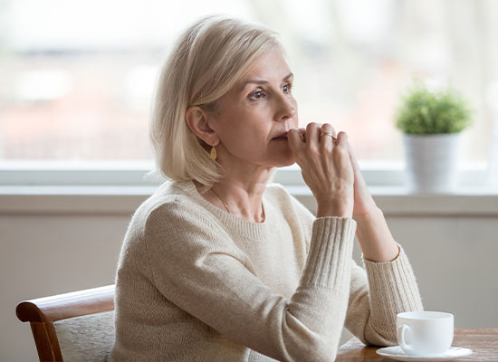 An elderly woman with short blonde hair and a light sweater sits at a table with a white cup, looking thoughtfully out the window. A potted plant is blurred in the background, creating a calm and reflective atmosphere, perhaps contemplating her estate planning under the new SECURE Act.