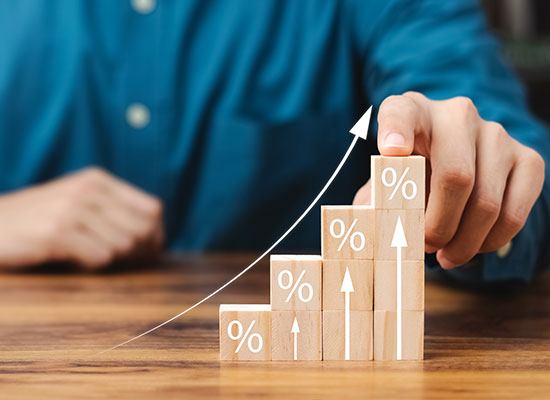 Person in a blue shirt points to the top block of a vertical staircase of wooden blocks on a table, each decorated with a percent sign and upward arrow, representing increasing growth or progress