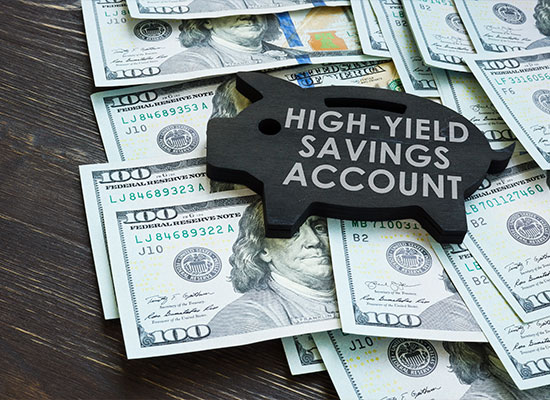 A wooden piggy bank shape with the words 'HIGH-YIELD SAVINGS ACCOUNT' rests on a pile of US $100 bills