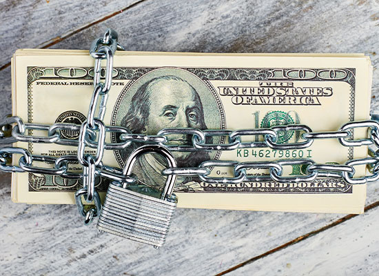 A stack of U.S. hundred-dollar bills, symbolizing investments, is wrapped with a chain secured by a padlock, placed on a wooden surface