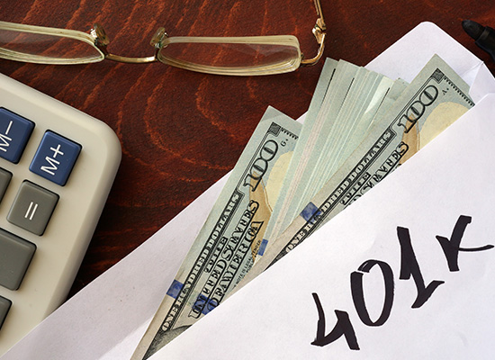 An envelope labeled '401k' is filled with $100 bills, placed next to a calculator and glasses on a wooden surface.
