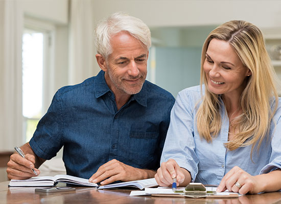 A senior man and a younger woman sit at a table, smiling as they look over documents. Both are casually dressed, with the man in a denim shirt and the woman in a light blue blouse. They appear to be engaged in financial planning, possibly discussing cost basis or studying paperwork.