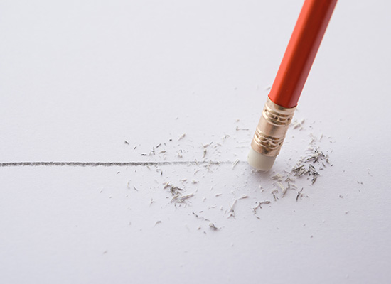 A red pencil eraser rubbing out a pencil line on white paper, leaving eraser shavings.