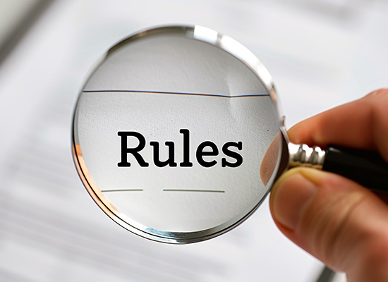 A magnifying glass held by a hand, focusing on the word "Rules" printed on a white surface.