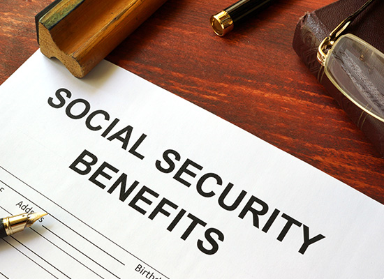 A close-up of a 'Social Security Benefits' application form with a pen, a pair of glasses, and a leather wallet on a wooden desk.