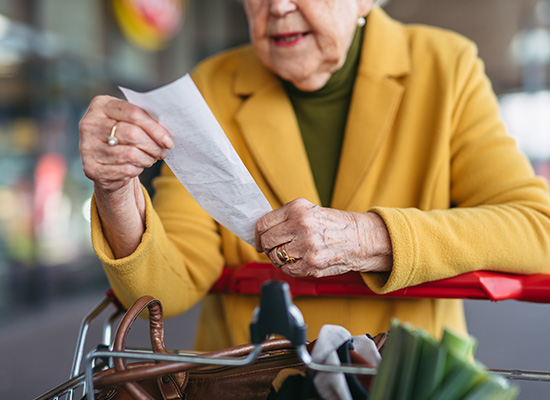 A senior woman in a yellow coat examines a receipt while standing with her shopping cart outside a store.
