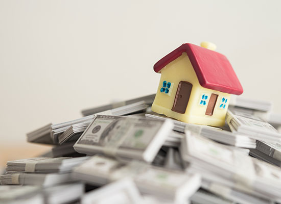 A small toy house resting on a pile of cash, representing the impact of inflation on purchasing power and the cost of living in real estate investments.