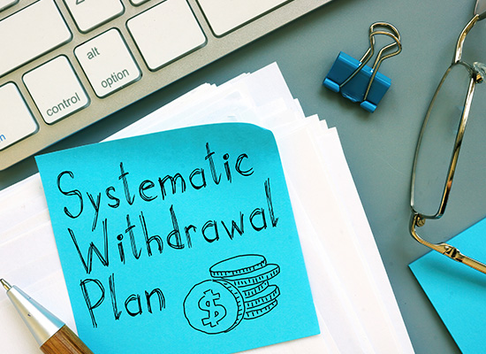 A sticky note with 'Systematic Withdrawal Plan' and a coin stack drawing sits on a desk with office supplies, depicting a retirement income strategy.