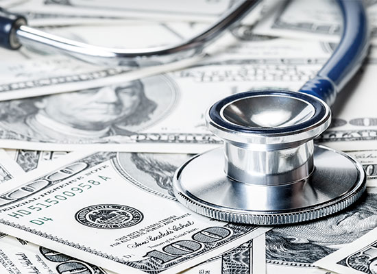 A stethoscope lies on top of multiple U.S. one hundred dollar bills, symbolizing the intersection of health care and financial costs.