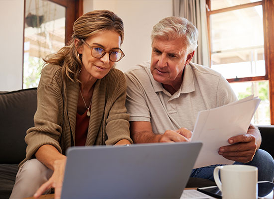 A middle-aged woman with glasses and a man with gray hair are sitting on a couch, focusing on a laptop screen.