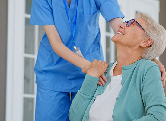 A healthcare worker in blue scrubs stands with their hand on the shoulder of a smiling elderly woman with short white hair and glasses, who is seated and wearing a light green cardigan