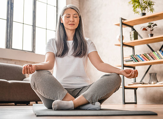 A middle-aged asian woman practicing meditation on a yoga mat in a serene living room, eyes closed and legs crossed in a lotus position.