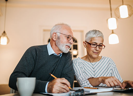 An elderly man and woman sit at a table, looking at documents related to social security together. The man holds a pencil while the woman points to a piece of paper. Both wear glasses and are focused on their task. A coffee mug is on the table, and warm lighting hangs above them.