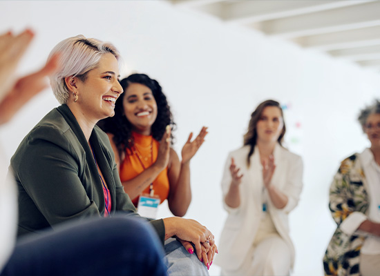 A diverse group of people sit in a bright room, attentively listening and clapping