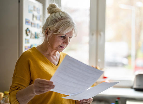 An older woman with gray hair in a bun stands in her kitchen, wearing a yellow sweater. She reviews retirement documents with a focused expression.