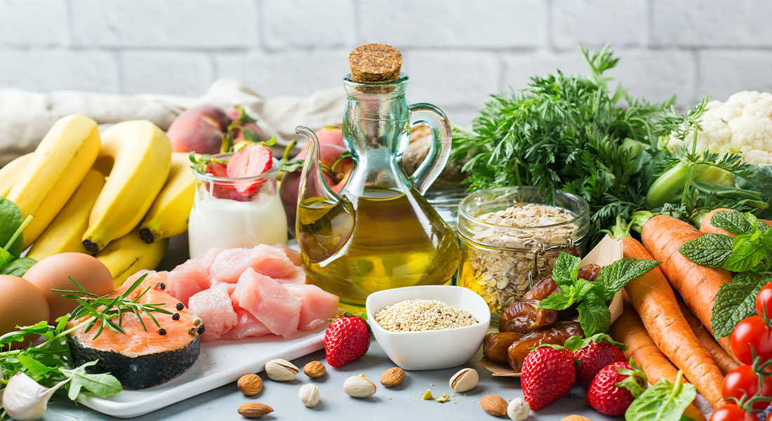 An array of healthy foods including fish, grains, fruits, carrots, bananas, eggs, olive oil, nuts, and leafy greens