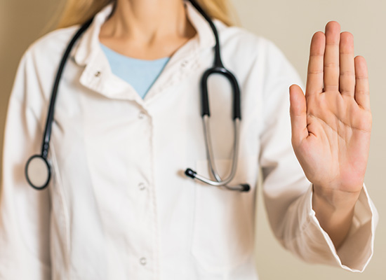 A healthcare professional wearing a lab coat with a stethoscope around the neck is holding up an open hand in a stop gesture.