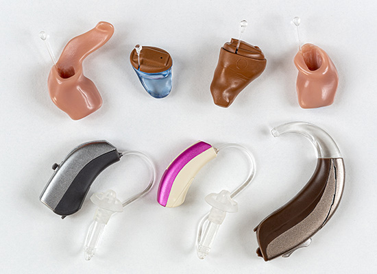 Various types of hearing aids in different colors and styles are displayed against a white background.