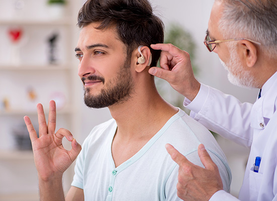 An audiologist is fitting a hearing aid on a young man who is signaling 'OK' with his hand, indicating a successful adjustment.