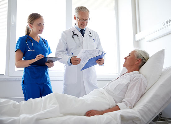A doctor and a nurse stand beside an elderly patient lying in a hospital bed. The doctor is holding a clipboard and discussing the patient's care, while the nurse takes notes on a notepad. Natural light floods the well-lit room through large windows in the background, emphasizing compassionate Medicare services