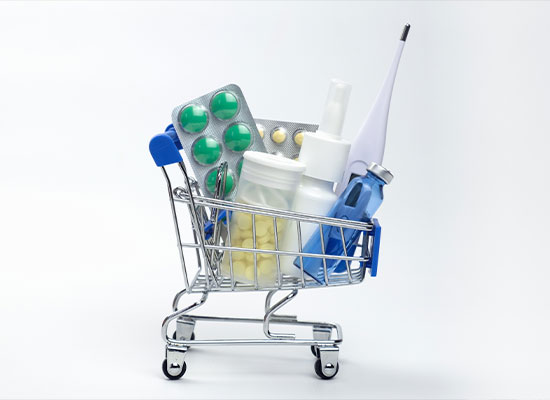 A miniature shopping cart filled with various Medicare medical supplies, including blister packs of pills, plastic bottles, and a digital thermometer, against a white background.