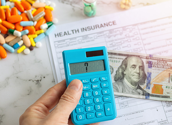 A person uses a blue calculator while reviewing health insurance documents, with pills and a $100 bill nearby, symbolizing the financial challenges and coverage gaps related to Medicare and out-of-pocket medical costs.