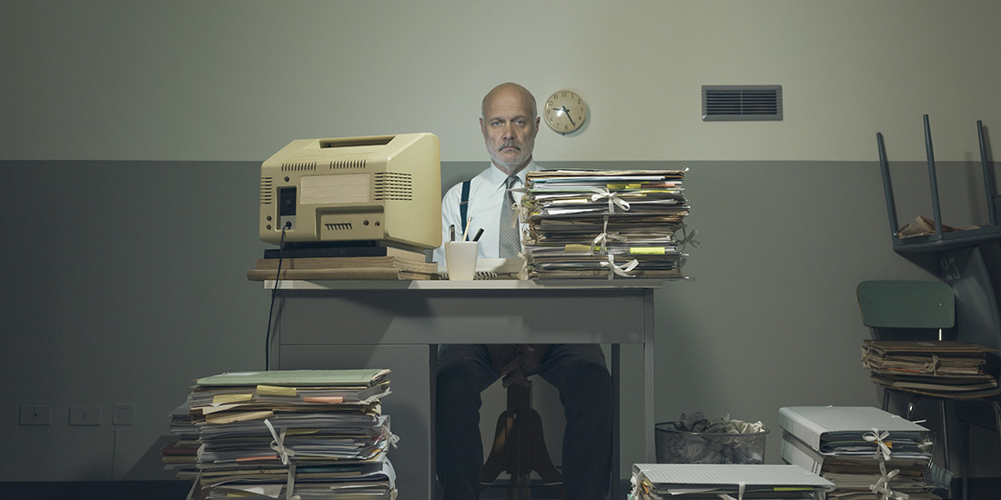 A senior man being overworked with stacks of files and documents on his desk and floor