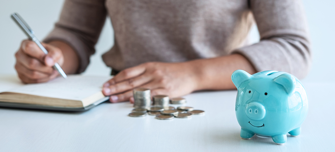 Woman filling out a ledger with her coins and piggy bank next to her.