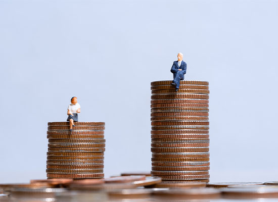 Miniature figures of a man and a woman sitting on two separate stacks of coins of varying heights, symbolizing financial inequality or savings.