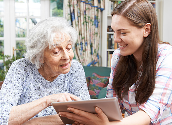 An elderly woman and a young woman smiling at each other while looking at a tablet together in a brightly lit living room.
