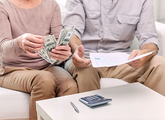 An elderly couple seated on a couch, engaged in retirement planning. The woman holds cash, while the man examines a document, with a calculator and pen on the table in front of them.