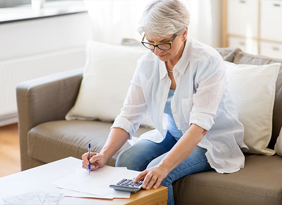 An older woman with short gray hair, wearing glasses and a casual shirt, is sitting at a coffee table using a calculator and writing notes, surrounded by papers related to retirement planning.