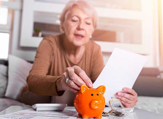 Elderly woman placing money into a piggy bank while reviewing documents, representing unique challenges in women's retirement.