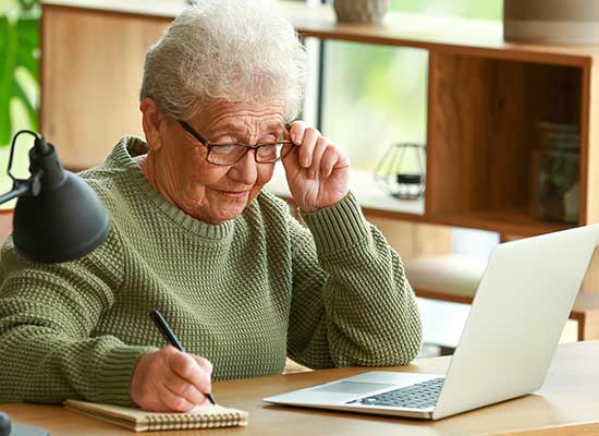 Senior woman taking notes while working on a laptop, showcasing women's retirement strategies and financial planning.
