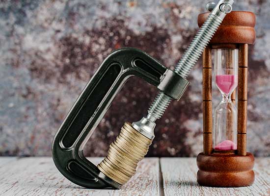A clamp squeezing a stack of coins next to an hourglass, illustrating the concept of retirees often underspending their savings to ensure financial security over time