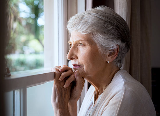An elderly woman with short gray hair gazes thoughtfully out of a window. She rests her hands on the windowsill, and natural light illuminates her face. She appears contemplative, perhaps pondering retirement planning, with a serene outdoor scene visible through the window behind her.