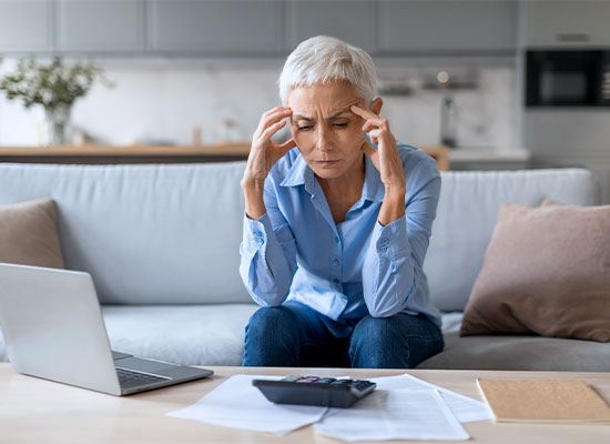 An elderly person with short gray hair sits on a couch in a modern living room, looking stressed. They are wearing a blue shirt and resting their head in their hands. A laptop, some papers, and a calculator—tools for retirement planning—are on the table in front of them.