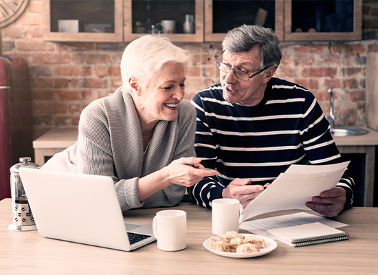 An elderly couple sits at a kitchen table, smiling and engaging with documents related to annuities. The man holds papers while the woman points at them. A laptop, coffee mugs, and a plate of pastries are on the table. The background features a brick wall and kitchen shelves.