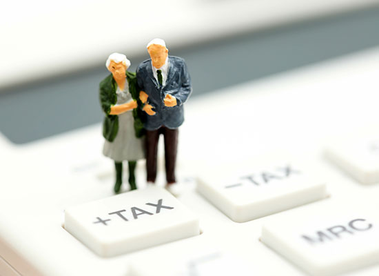 A miniature figurine of an elderly couple stands on a large calculator, specifically on the buttons labeled +TAX' and '-TAX.'