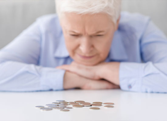 An elderly person with short white hair, wearing a light blue shirt, rests their head on their arms while looking intently at a small pile of coins spread out on a white surface, contemplating the impact of social security.