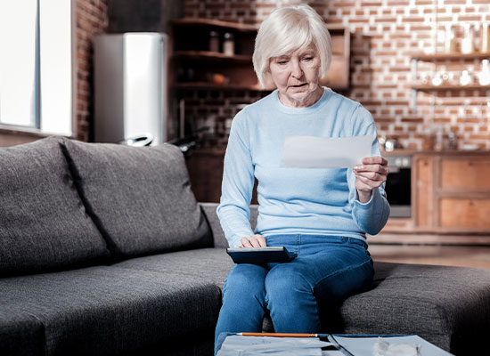 An elderly woman with short white hair sits on a gray couch in a cozy, modern living room. She is holding a piece of paper in one hand and a calculator in the other, looking at the paper with a focused expression. A table with social security documents is in front of her.
