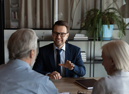 A smiling man in a suit and glasses sits at a desk engaging in conversation about social security with an older couple. The couple is seated with their backs to the camera. Shelves filled with books and a large potted plant adorn the background.