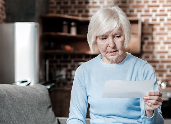 An elderly woman with white hair wearing a light blue sweater is sitting on a gray couch in a kitchen area, holding and looking at a piece of paper with a serious expression.