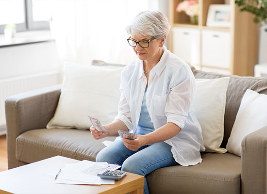 An elderly woman with short gray hair and glasses sits on a sofa, holding currency notes in one hand and a pen in the other.